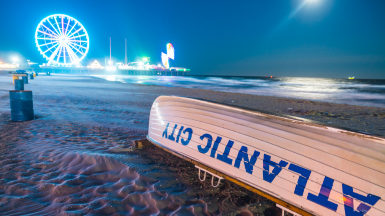An Atlantic City Lifeboat sits upside down on the beach and shoreline on a moonlit night with Steel Pier rides and Ferris Wheel lit up in background.